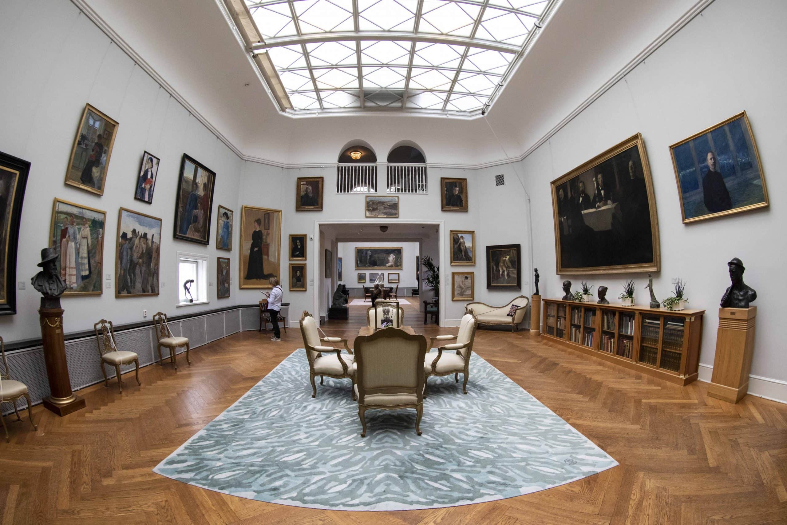 Visitors viewing paintings in a spacious art gallery with a skylight and classic furniture.