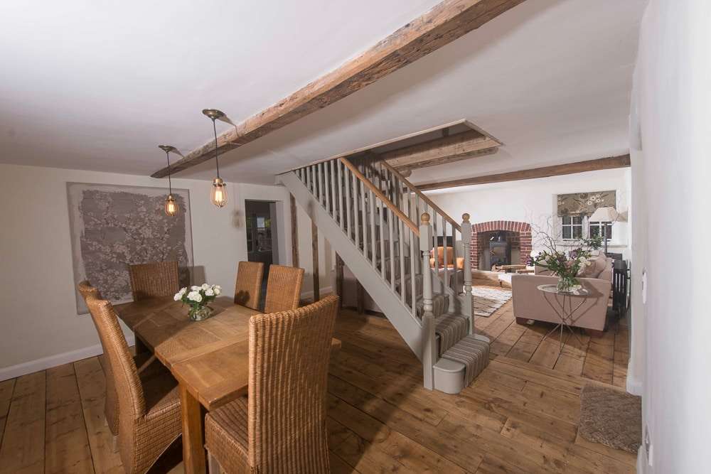 Interior view of a rustic home showing wooden dining set, exposed beams, stairway, and living area with a fireplace in the background.