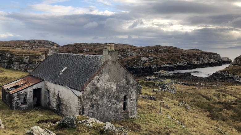 An old, weathered cottage with a damaged extension overlooking a rocky coastline under a cloudy sky.