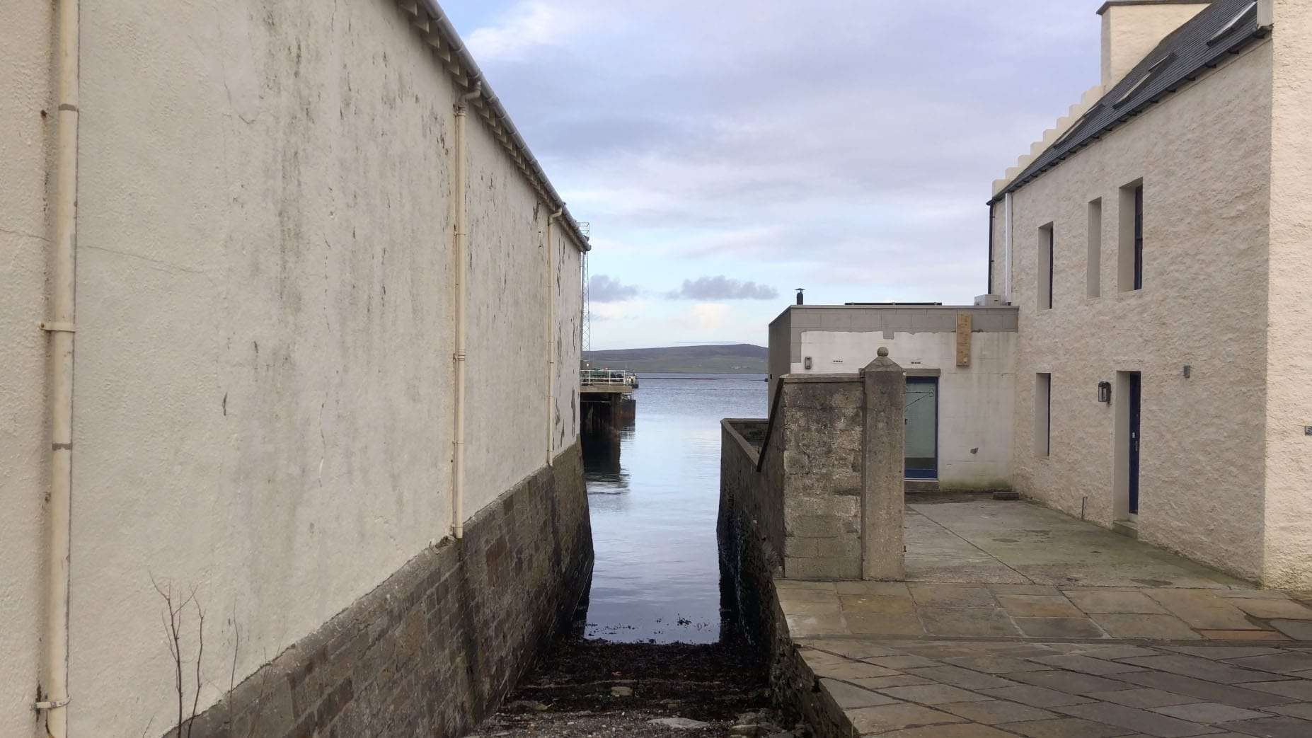 View of a narrow alley leading to a body of water with white buildings on either side under a cloudy sky.