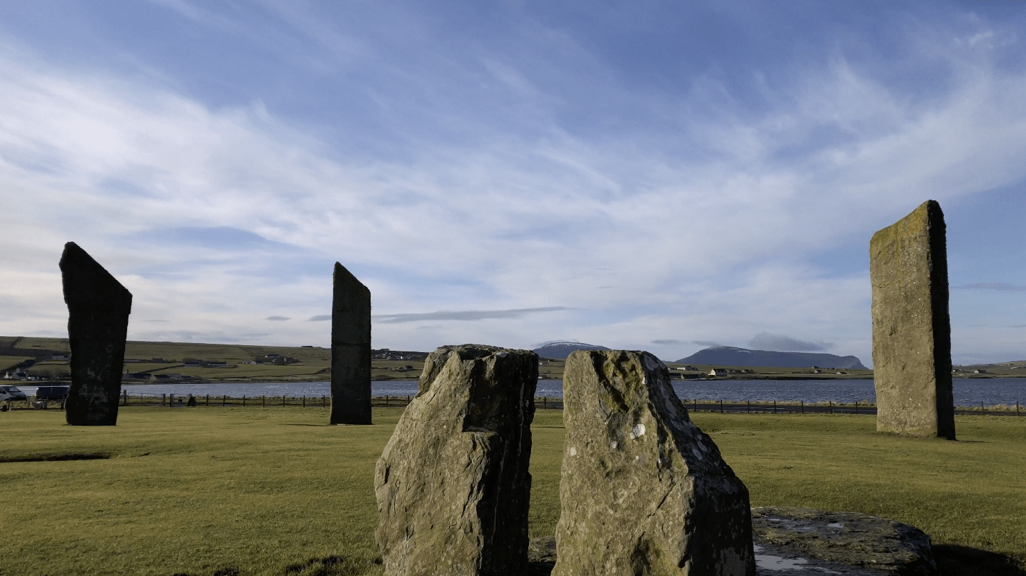 Several tall, ancient standing stones erected in a grassy field with a hilly and coastal backdrop under a blue sky with scattered clouds.