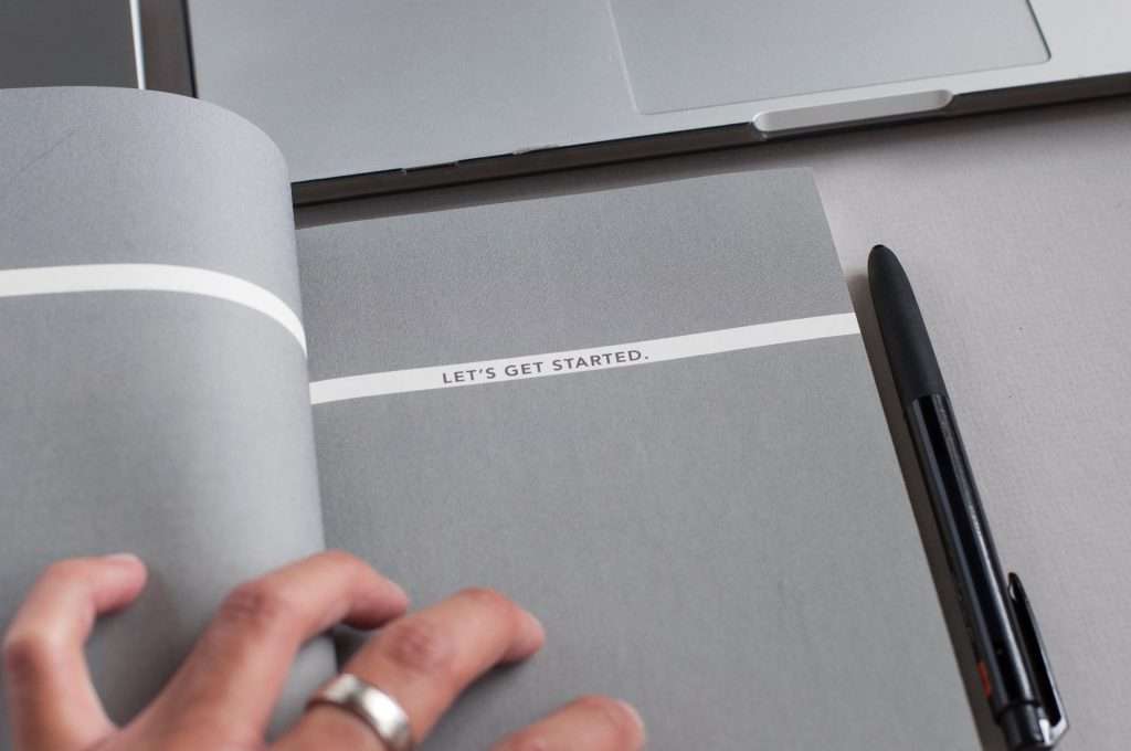 A person's hand turning a page of a notebook with "LET'S GET STARTED." written on it, beside a closed laptop and a pen.