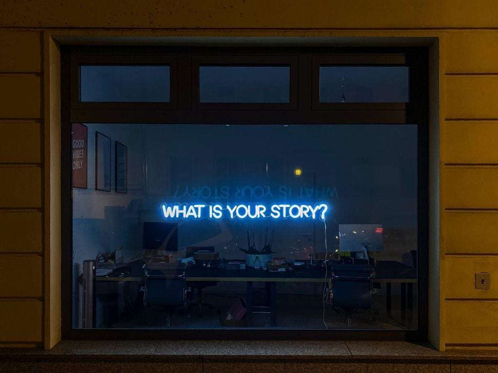 Neon sign with the question "WHAT IS YOUR STORY?" reflected in an office window at night, with visible desks and chairs inside.