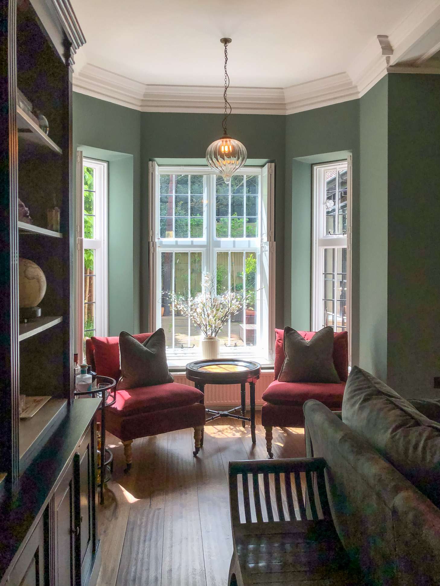 Interior view of a stylish sitting area with green walls, a pendant light, and red accent chairs by a window with garden view.