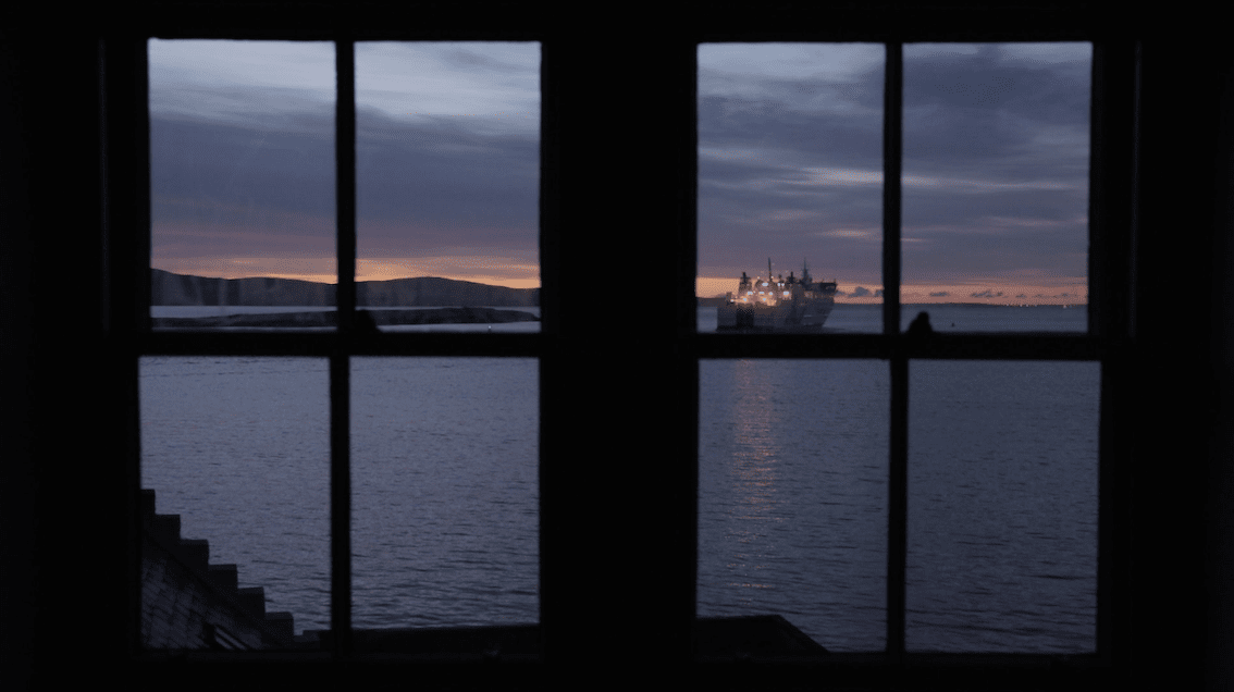 Ocean view at dusk featuring a ship on the horizon, seen through a dark window frame with subtle sunset colors in the sky.