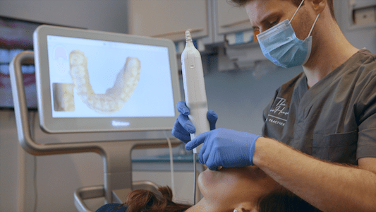 A dentist wearing a mask and gloves using a digital scanning device on a patient with a dental impression displayed on the screen in the background.