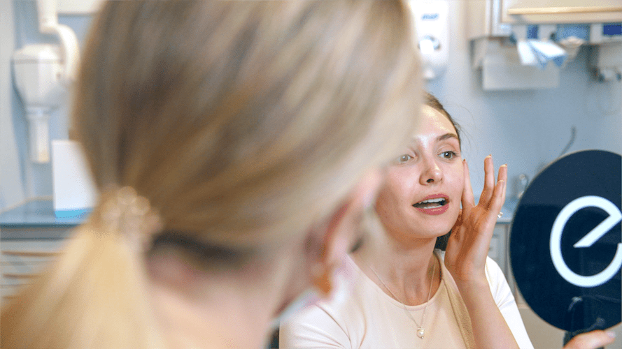 A woman examines her face in a mirror during a dermatology appointment.