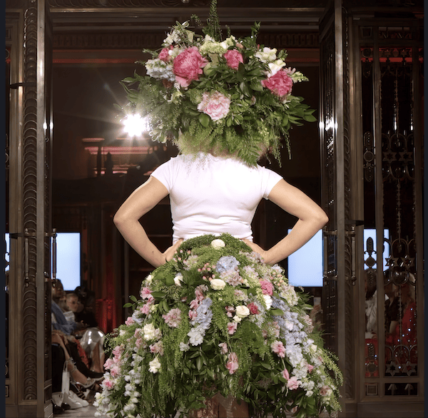 Person wearing an elaborate floral arrangement on their head and as a skirt, posing in a room with an audience in the background.