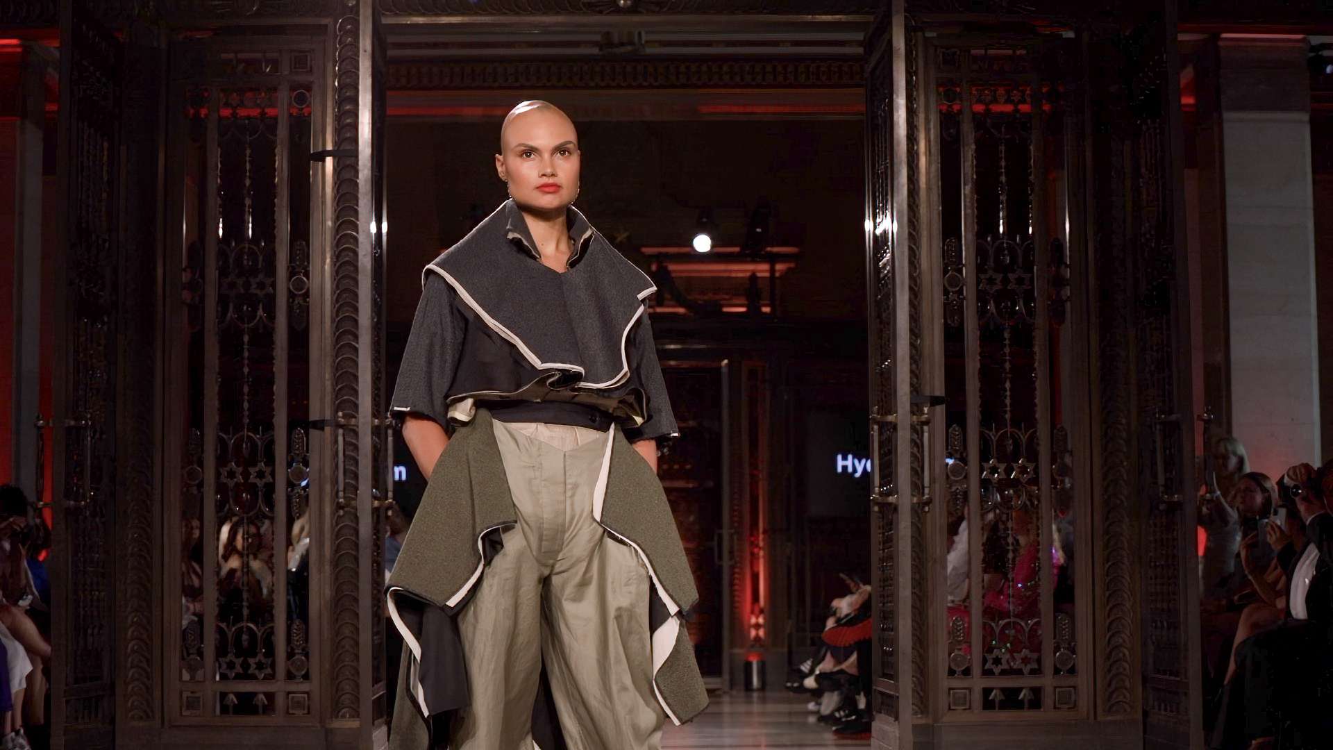 A model walking on the runway during a fashion show, showcasing an avant-garde outfit with spectators in the background.