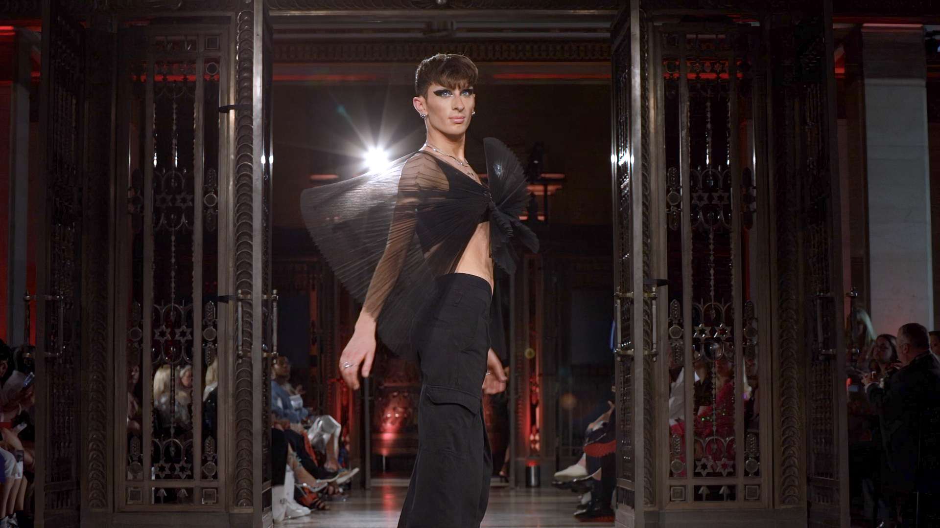 A model striking a pose on the catwalk at a fashion show event, wearing a black avant-garde outfit with a striking sheer winged accessory.
