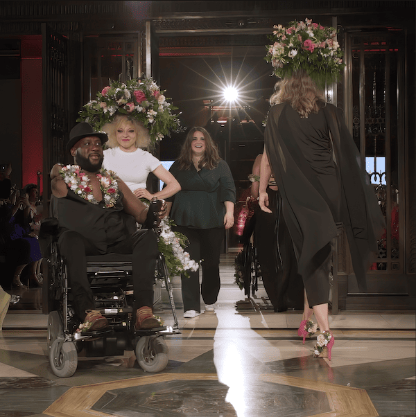 A diverse group of models on a fashion show runway, including two individuals in wheelchairs adorned with floral arrangements, with spectators in the background.