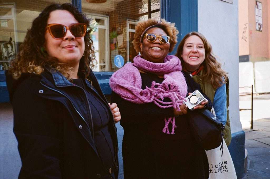 Three happy women of various ethnicities smiling and posing together on a city street, one holding a camera.
