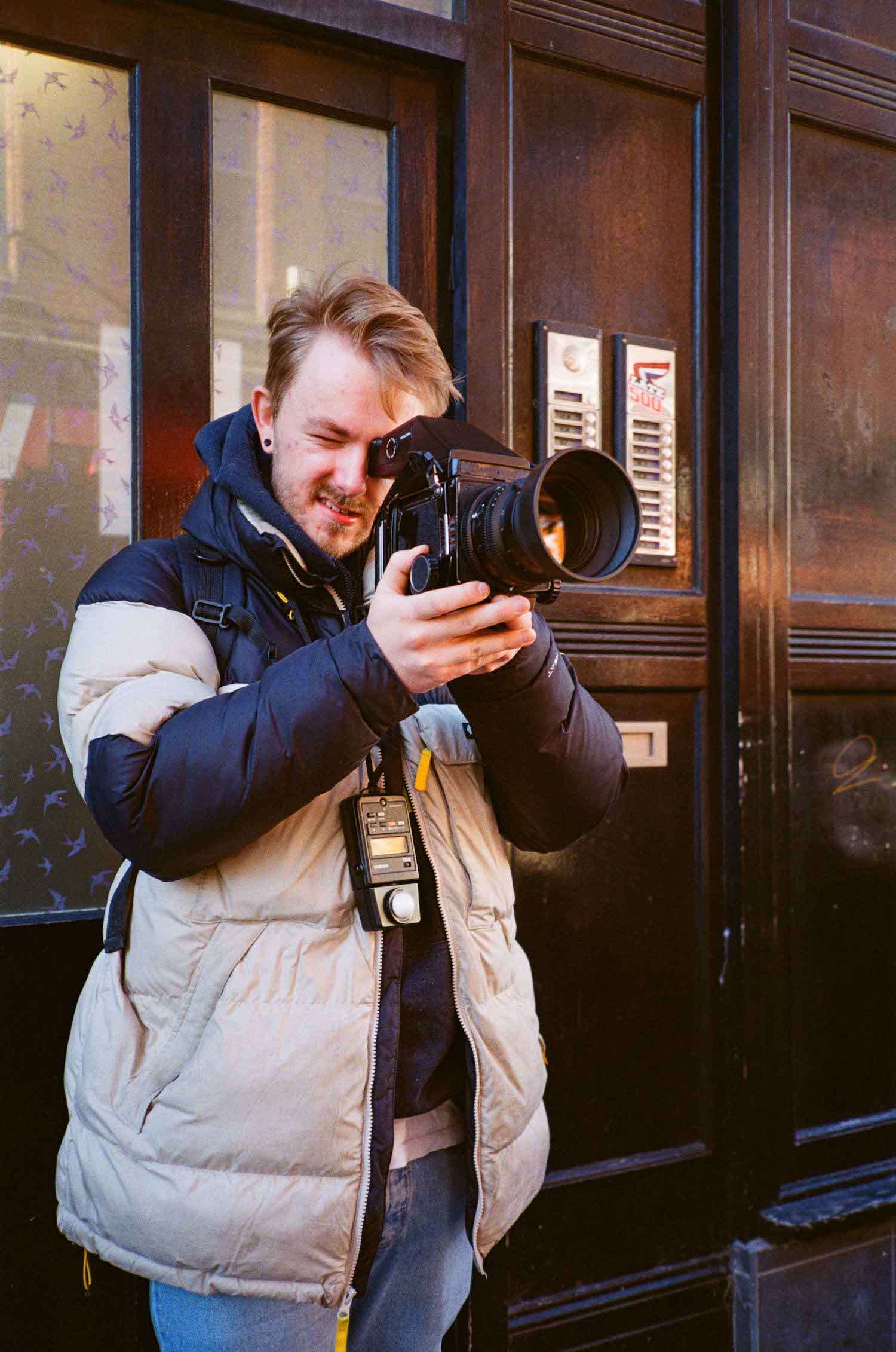 A person taking a photograph with a professional camera on a city street, wearing a light jacket and carrying a light meter.