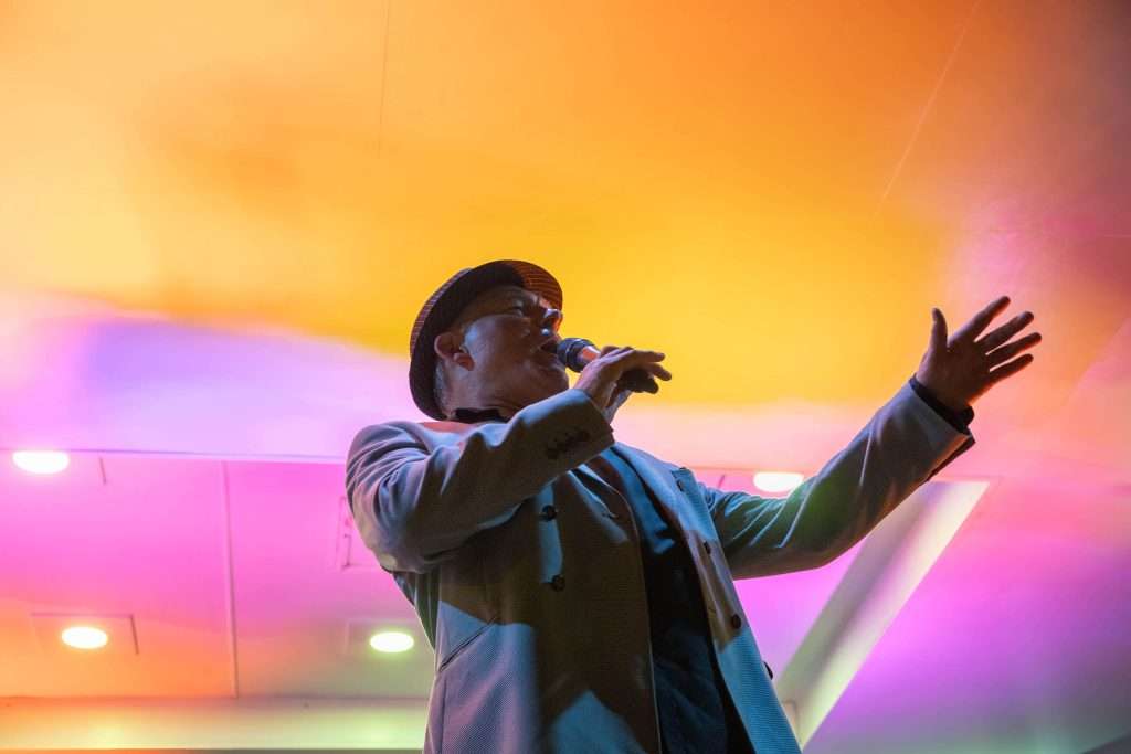 A person singing on stage with a hat, gesturing emotionally under a vibrant gradient of stage lights.