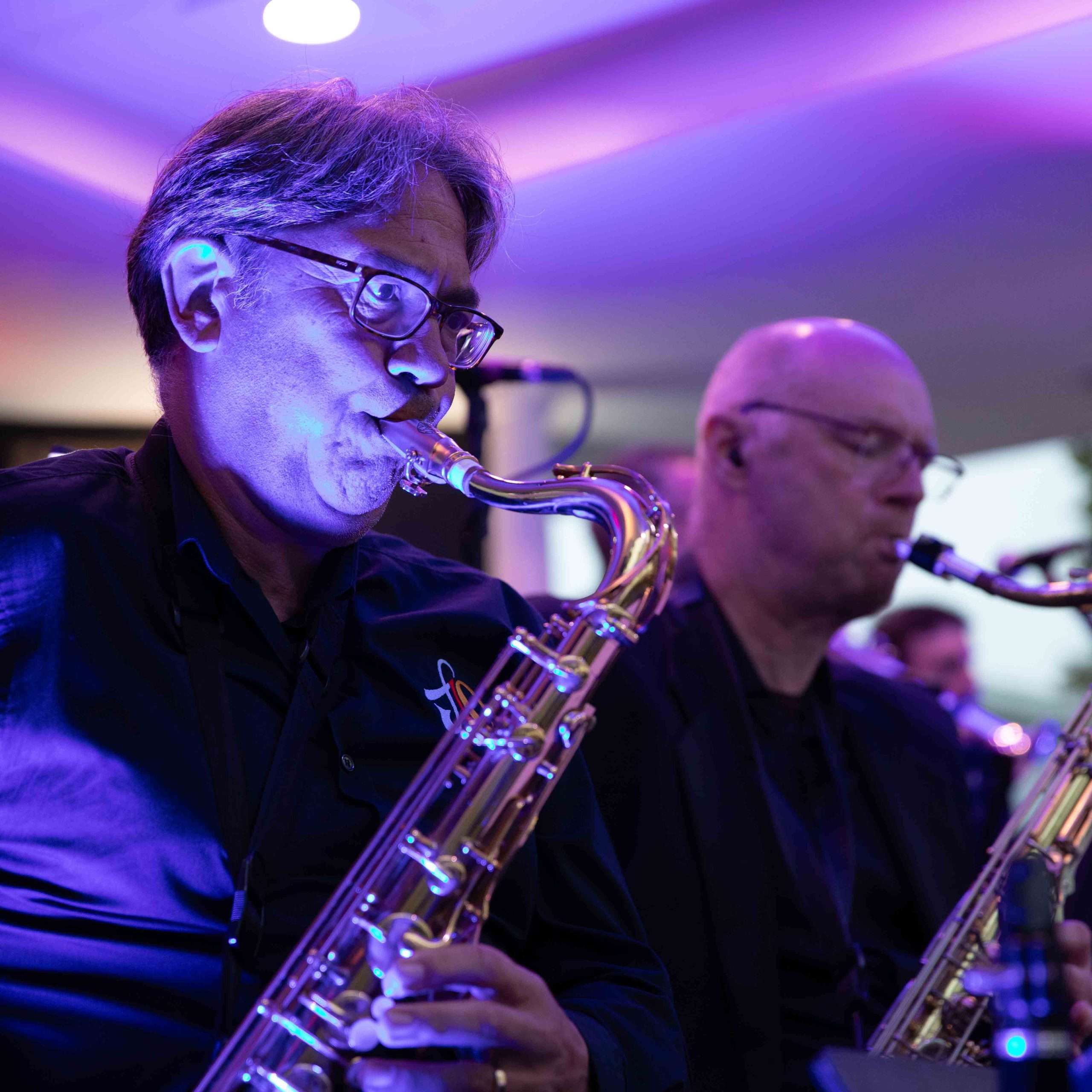 Two saxophonists playing in a band with a colorful stage lighting setup.