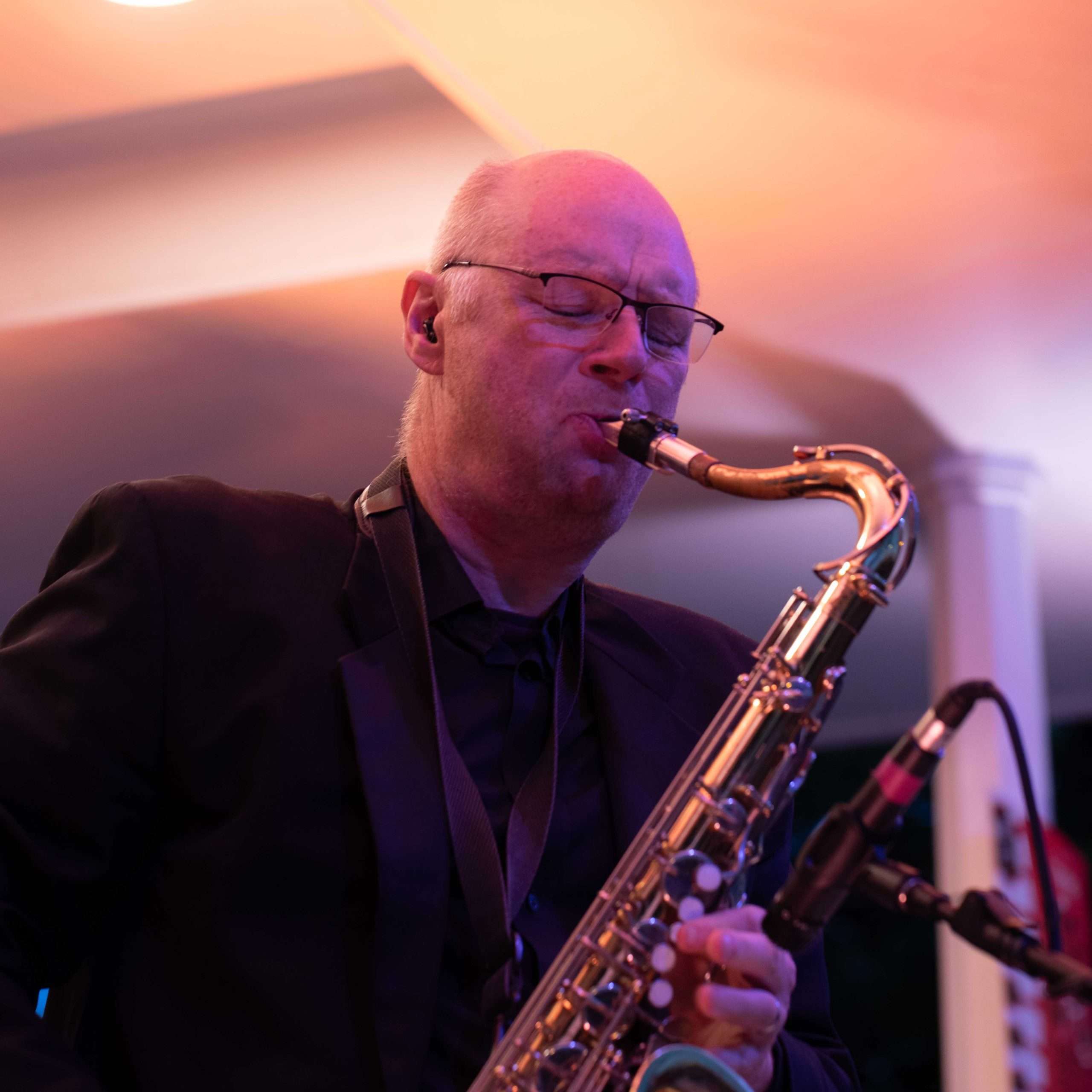 A man playing the saxophone during a live performance, with soft lighting in the background.