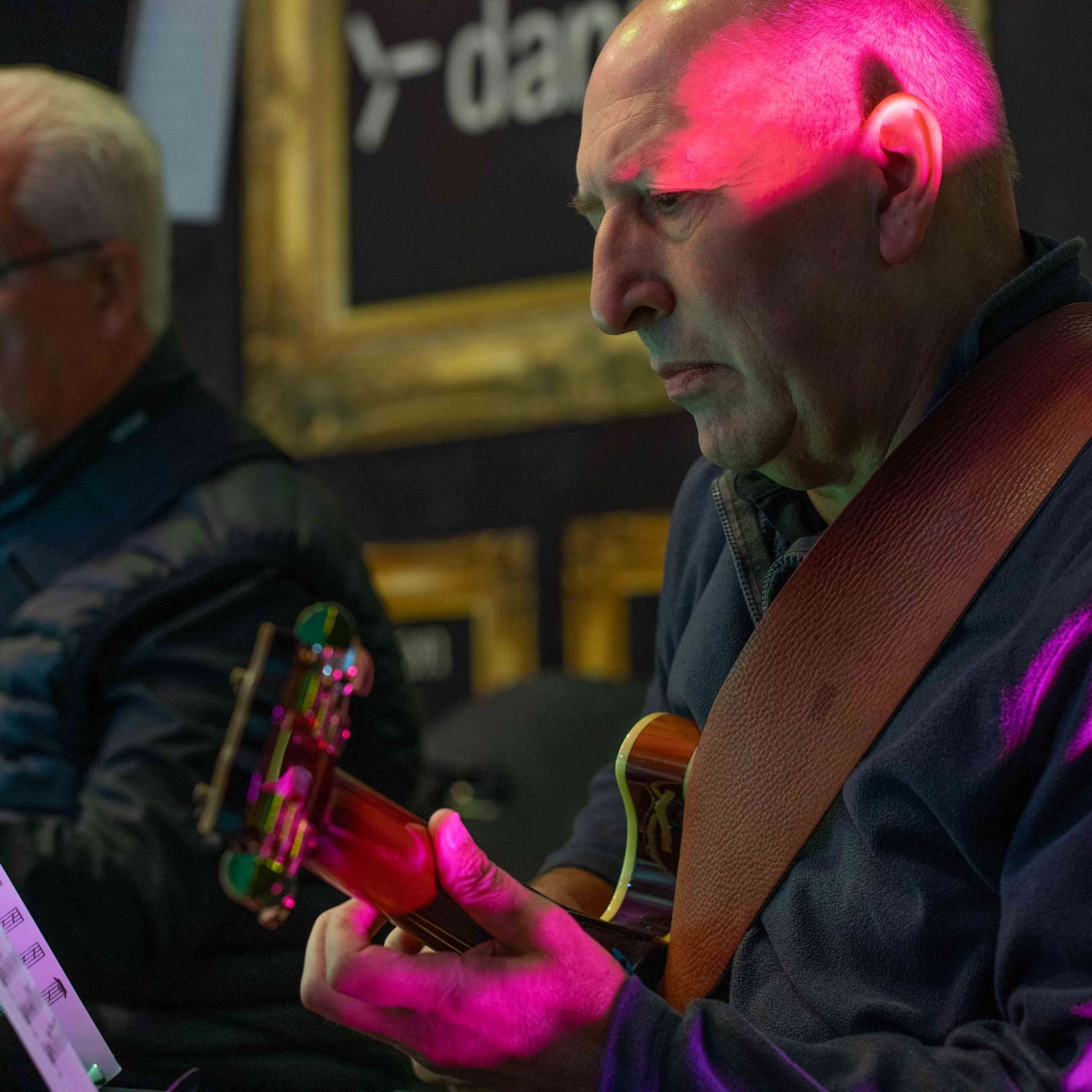 A musician focused on playing his acoustic guitar with a red stage light shining on him, another person in the background.