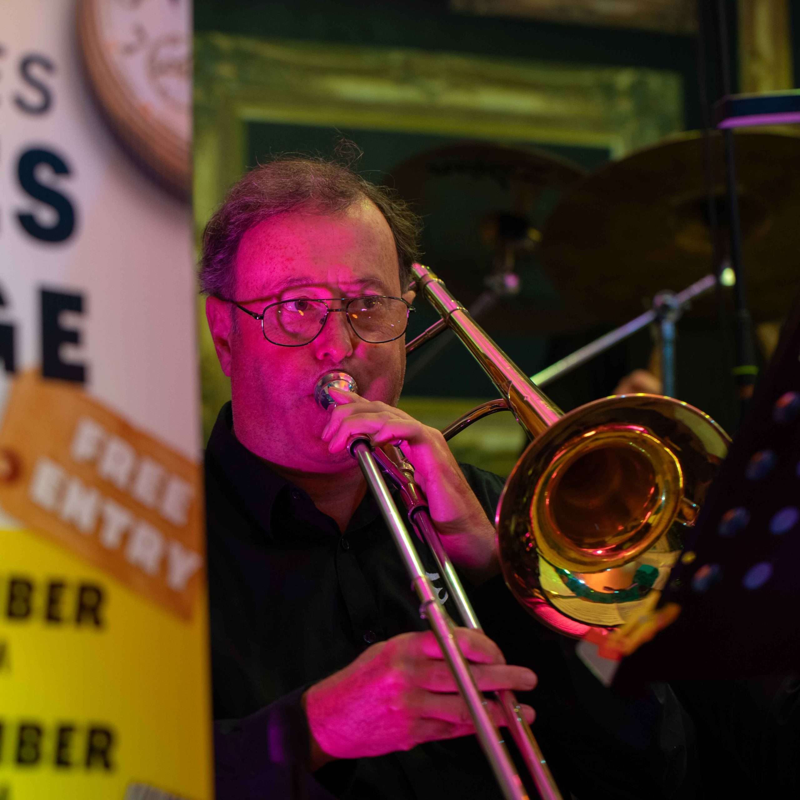 Man playing a trombone on stage with music stand and drum set in the background, at a venue offering free entry.