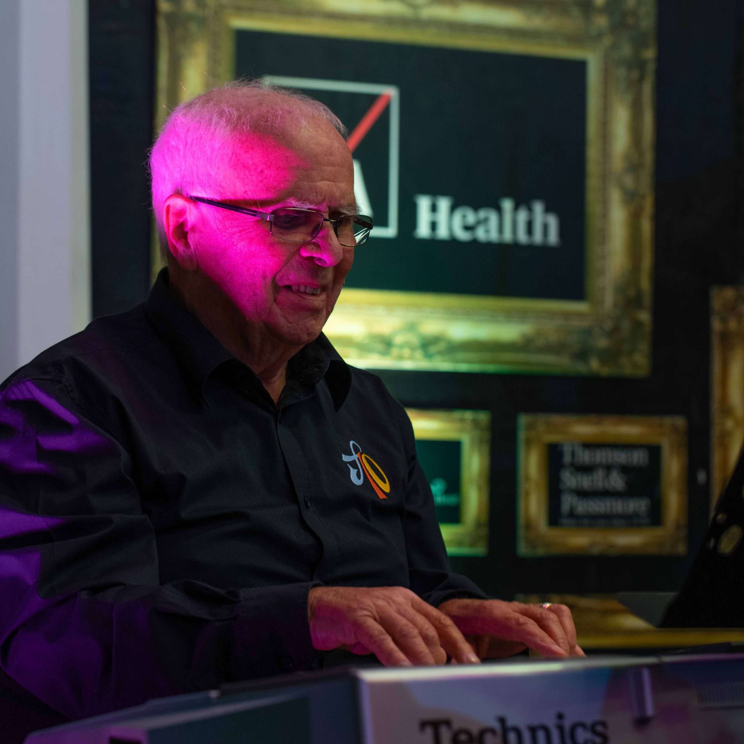 An older man with glasses playing a Technics electronic keyboard, lit by pink stage lighting with a sign reading "Health" in the background.