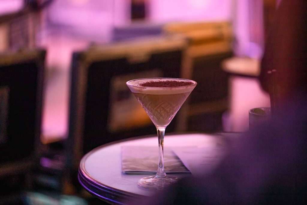 A martini glass with a chocolate garnished drink on a bar table with a purple hued background.