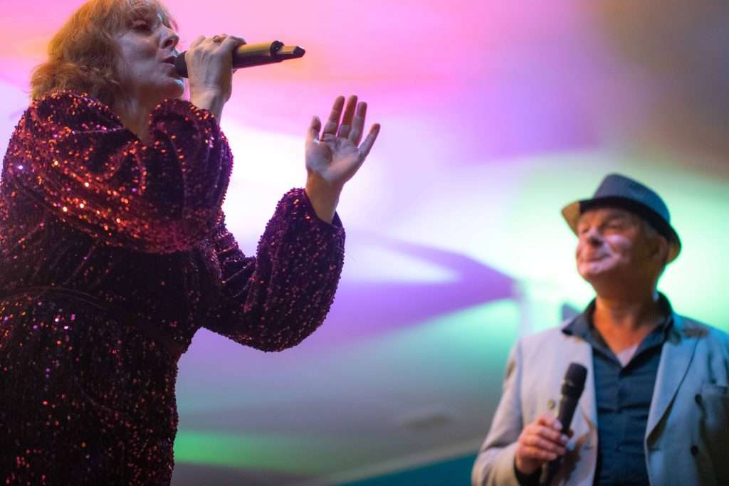 Two singers performing on stage, with the woman in a sparkly outfit singing into a microphone and the man in a hat holding a microphone by his side, both illuminated by vibrant stage lighting.