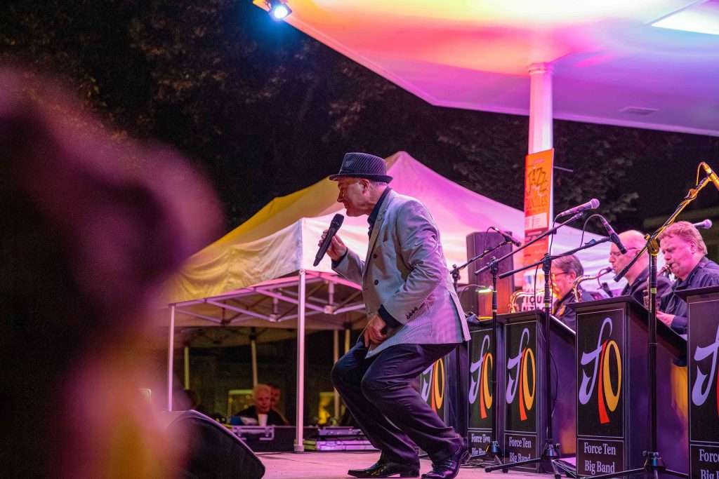 A singer performing on stage with the Force Ten Big Band at an outdoor evening event, colorful stage lighting with musicians in the background.