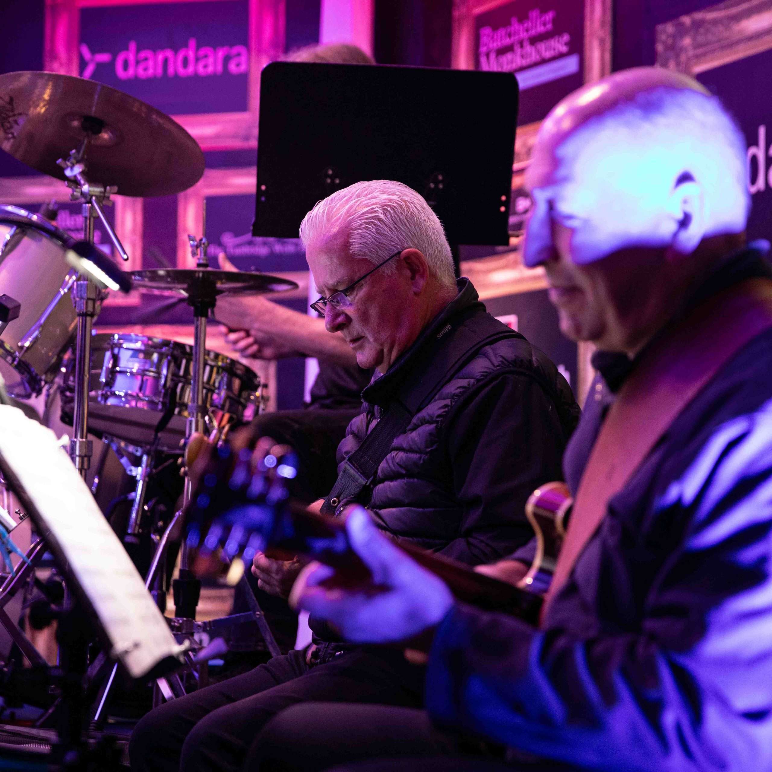 Two musicians playing instruments with a drummer in the background on stage during a live performance.