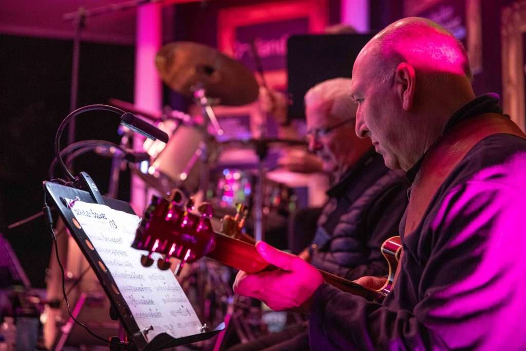 A guitarist and drummer performing on stage with a focus on the guitarist reading sheet music.