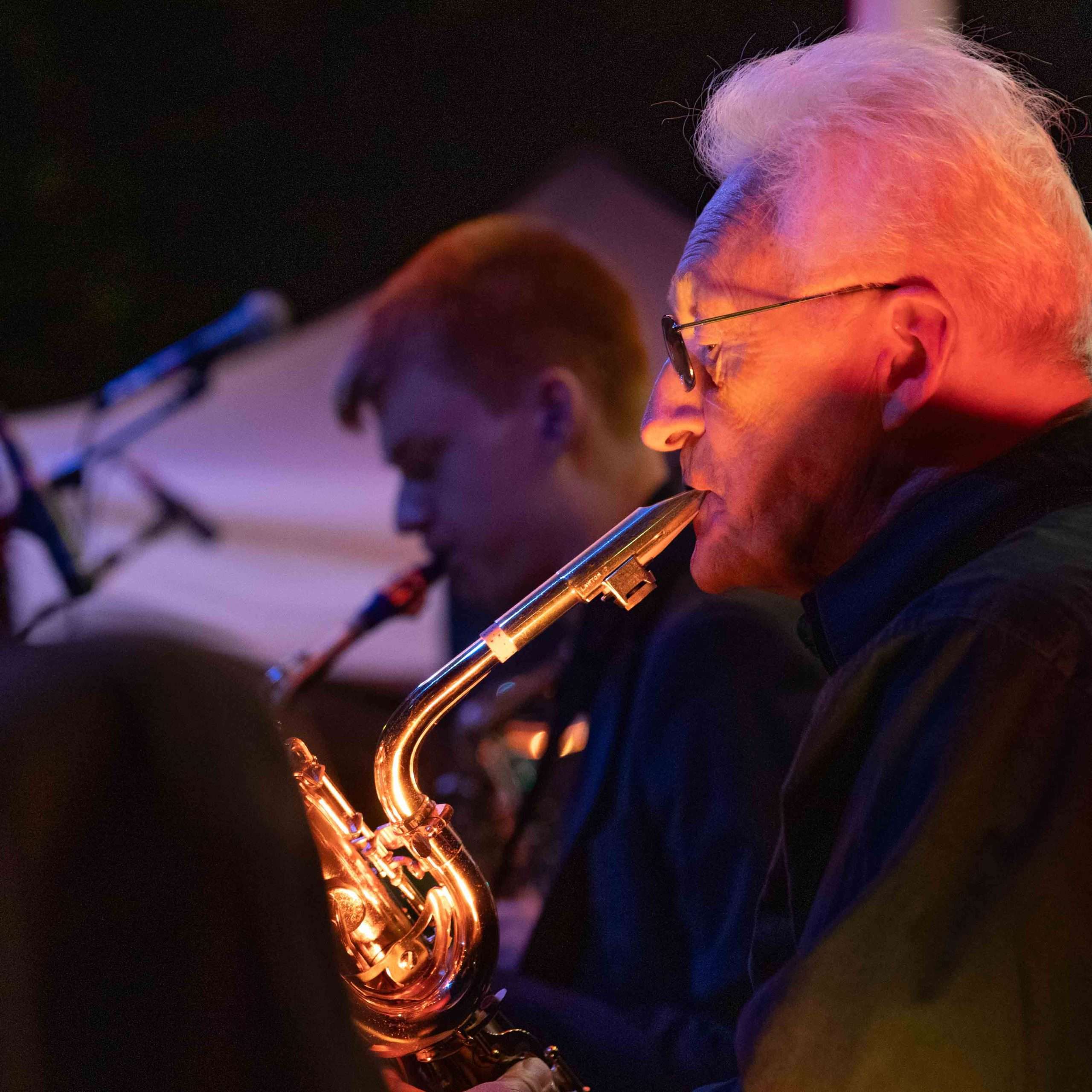 Two musicians playing saxophones on stage illuminated by warm stage lighting.