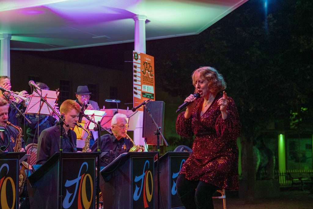 A woman in a sparkling dress singing with a microphone, accompanied by a live jazz band under colored stage lighting.