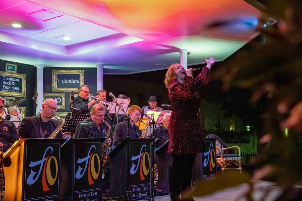 A female singer performing with a jazz big band on an outdoor stage with colorful lighting, surrounded by musicians with saxophones, trumpets, and music stands displaying the band's logo.