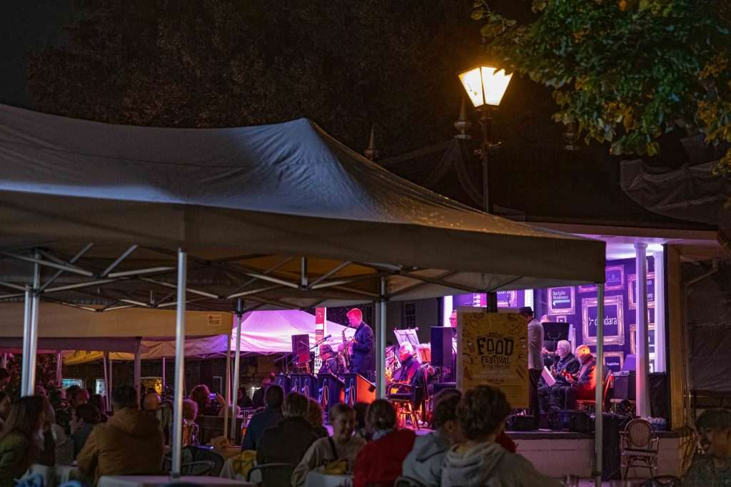 People enjoying a live performance at an outdoor food festival at night, with a band on stage and a streetlamp illuminating the area.