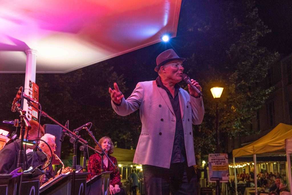 A man in a hat singing with a microphone in front of a live band on a stage with colorful lighting at an outdoor evening event
