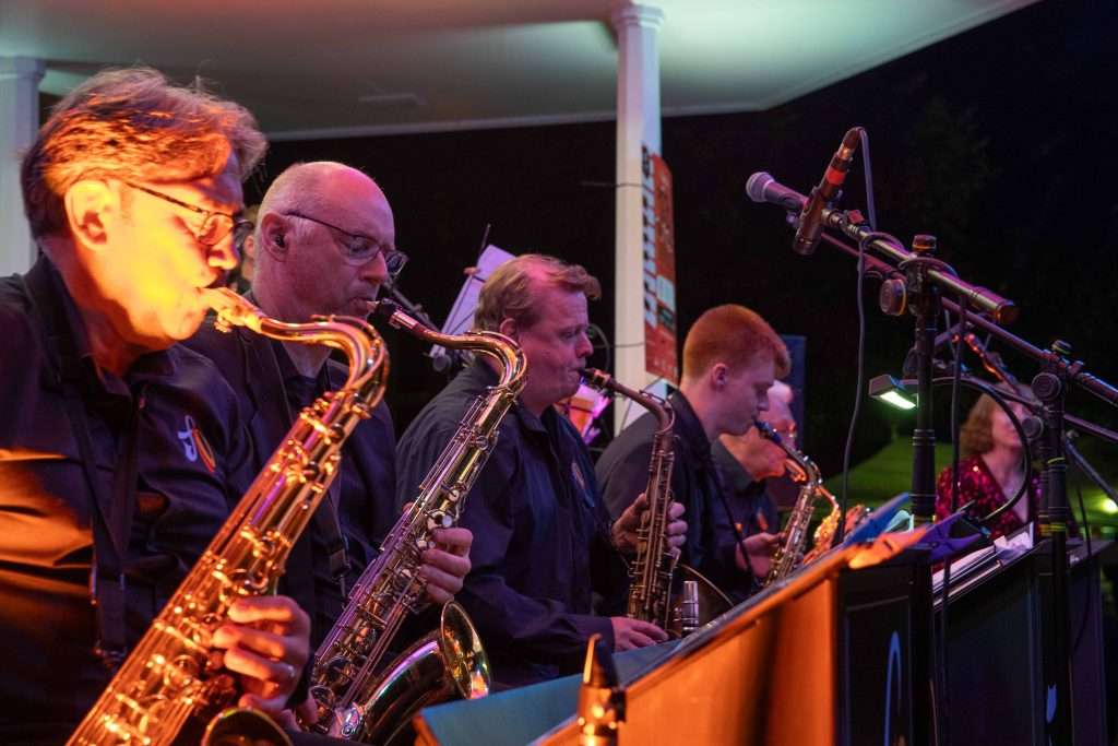 A group of saxophonists playing their instruments on stage during an evening outdoor concert with microphones and music stands