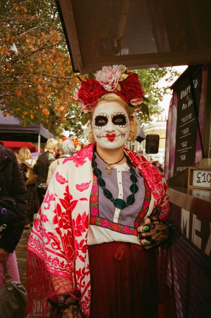 Person dressed in traditional Day of the Dead attire with skull makeup and a floral headdress standing at an outdoor market.
