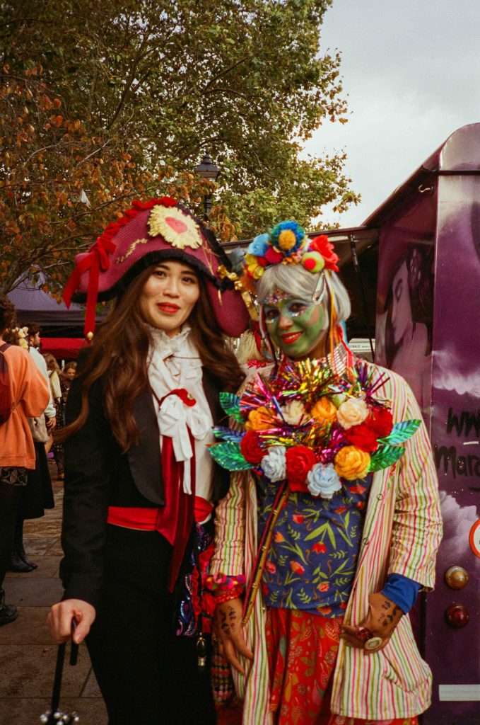 Two people dressed in vibrant and elaborate costumes at an outdoor event, with one in traditional pirate attire and the other in a colorful, whimsical outfit with face paint.