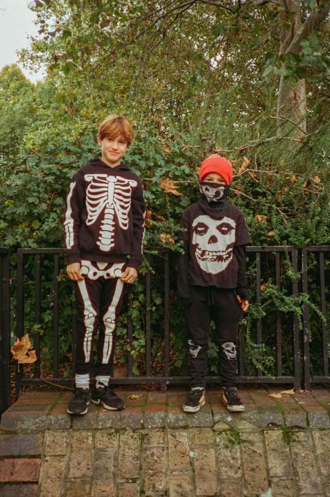Two children dressed in skeleton-themed costumes standing in front of a fence with greenery in the background.
