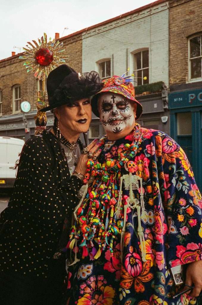 Two individuals dressed in elaborate Day of the Dead costumes, one with a skull face paint and colorful attire, standing on a street.