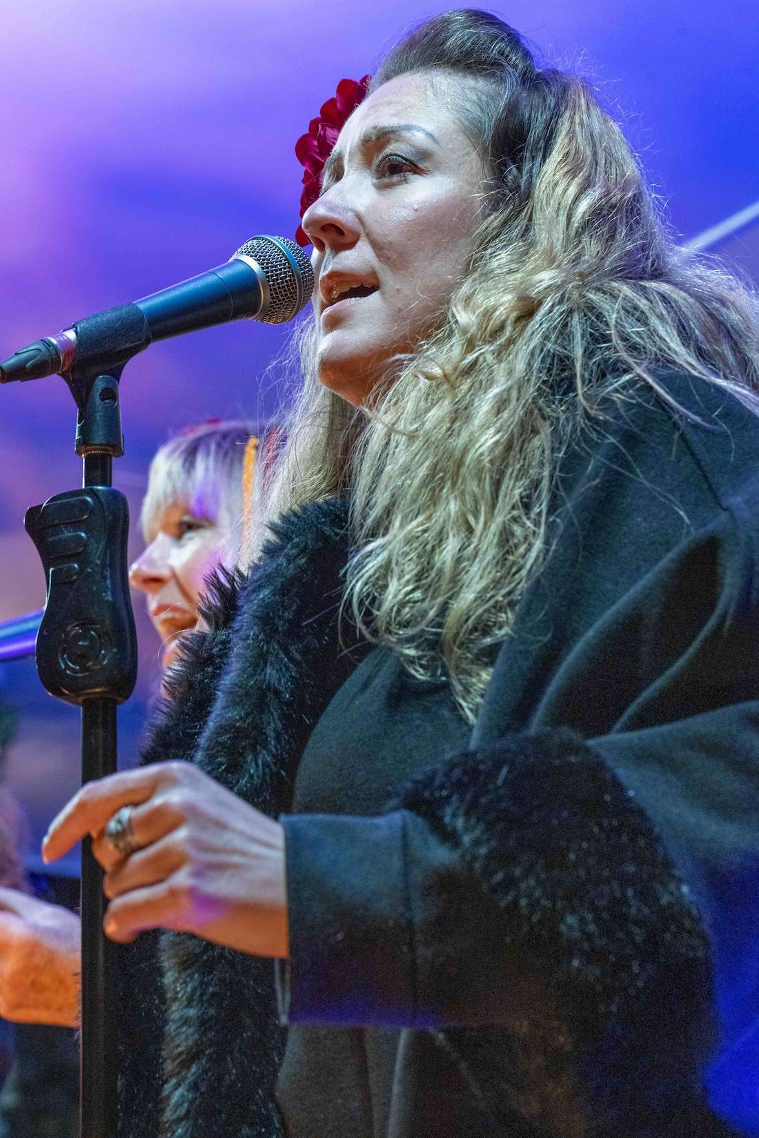 A woman singing into a microphone with a red flower in her hair, with another person in the background.