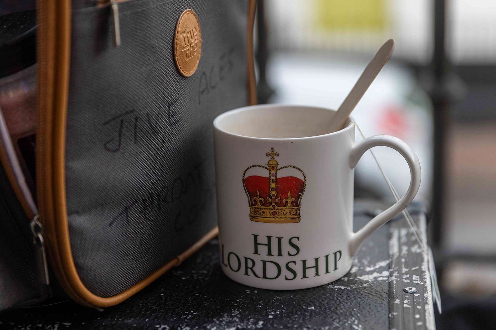 A close-up of a white mug with "HIS LORDSHIP" printed on it, next to a bag with inscriptions, on a dark surface with raindrops.
