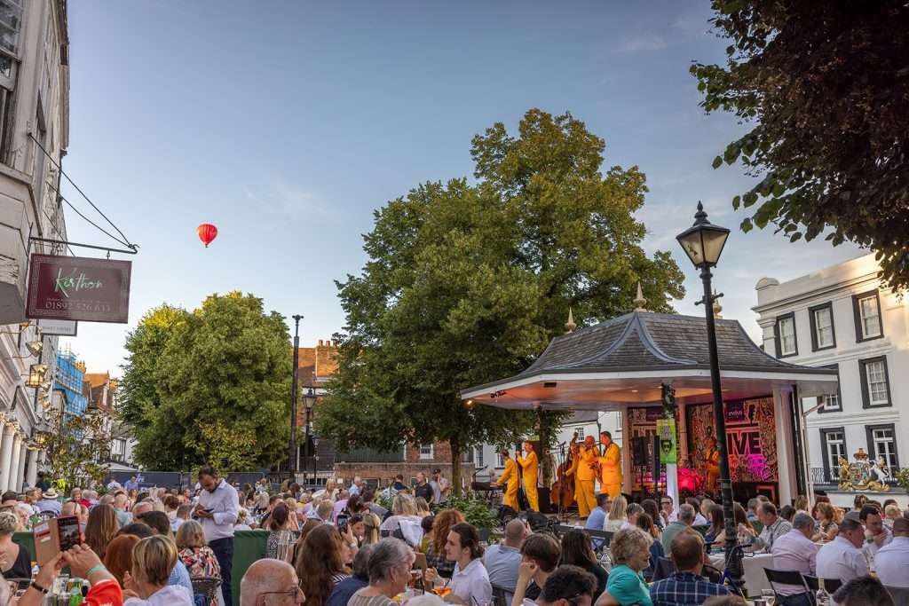 Crowd of people enjoying an outdoor live music performance under a gazebos with a red hot air balloon floating in the blue sky in the background.