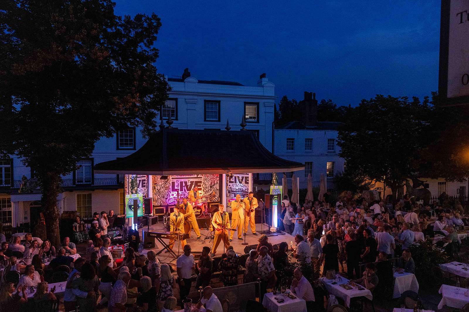 A lively crowd enjoys an evening outdoor concert with a band performing on stage, surrounded by historic buildings under a twilight sky.