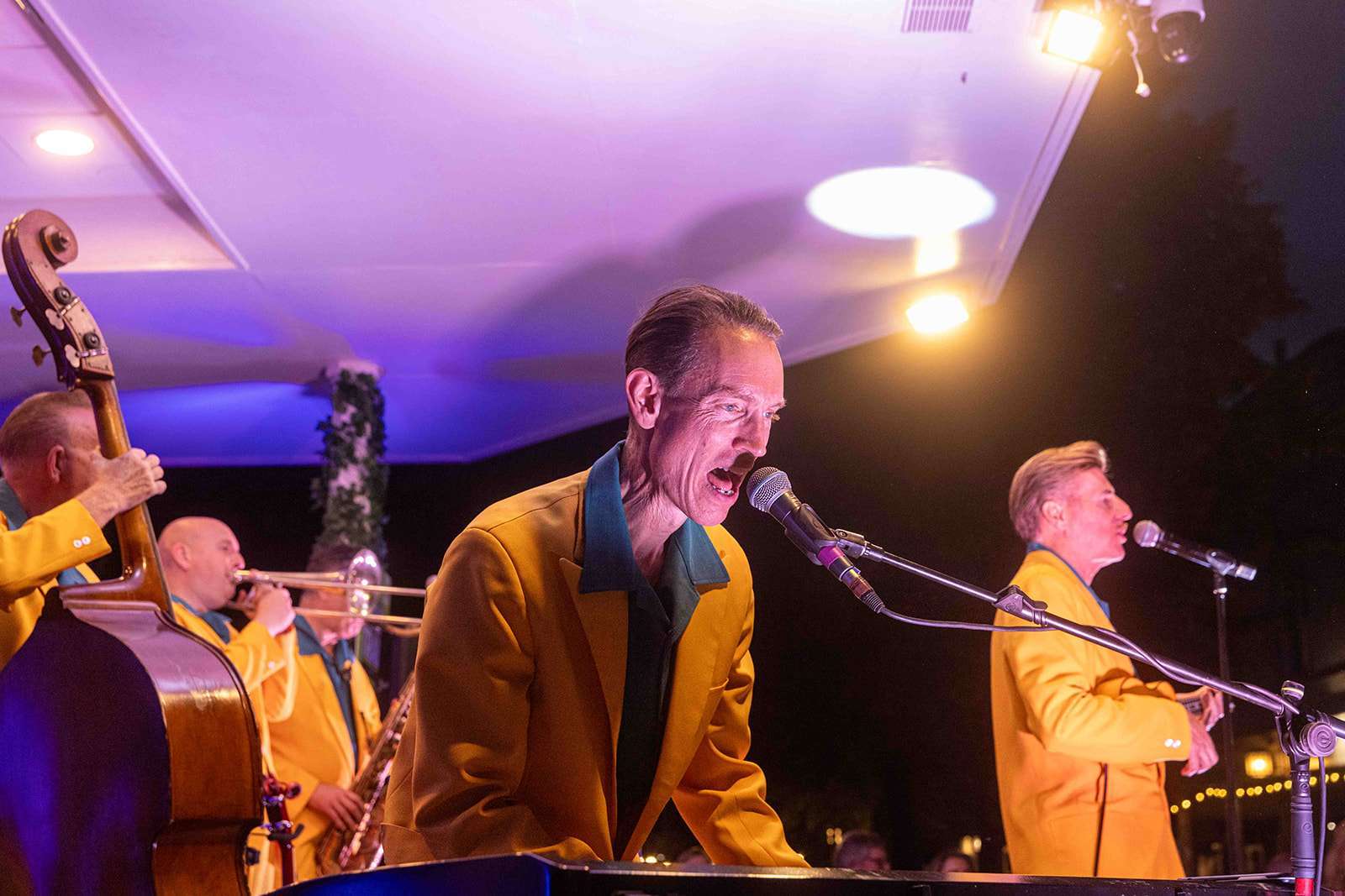 A jazz band performing live with a singer at a microphone and instrumentalists playing a double bass and a trombone with stage lights in the background