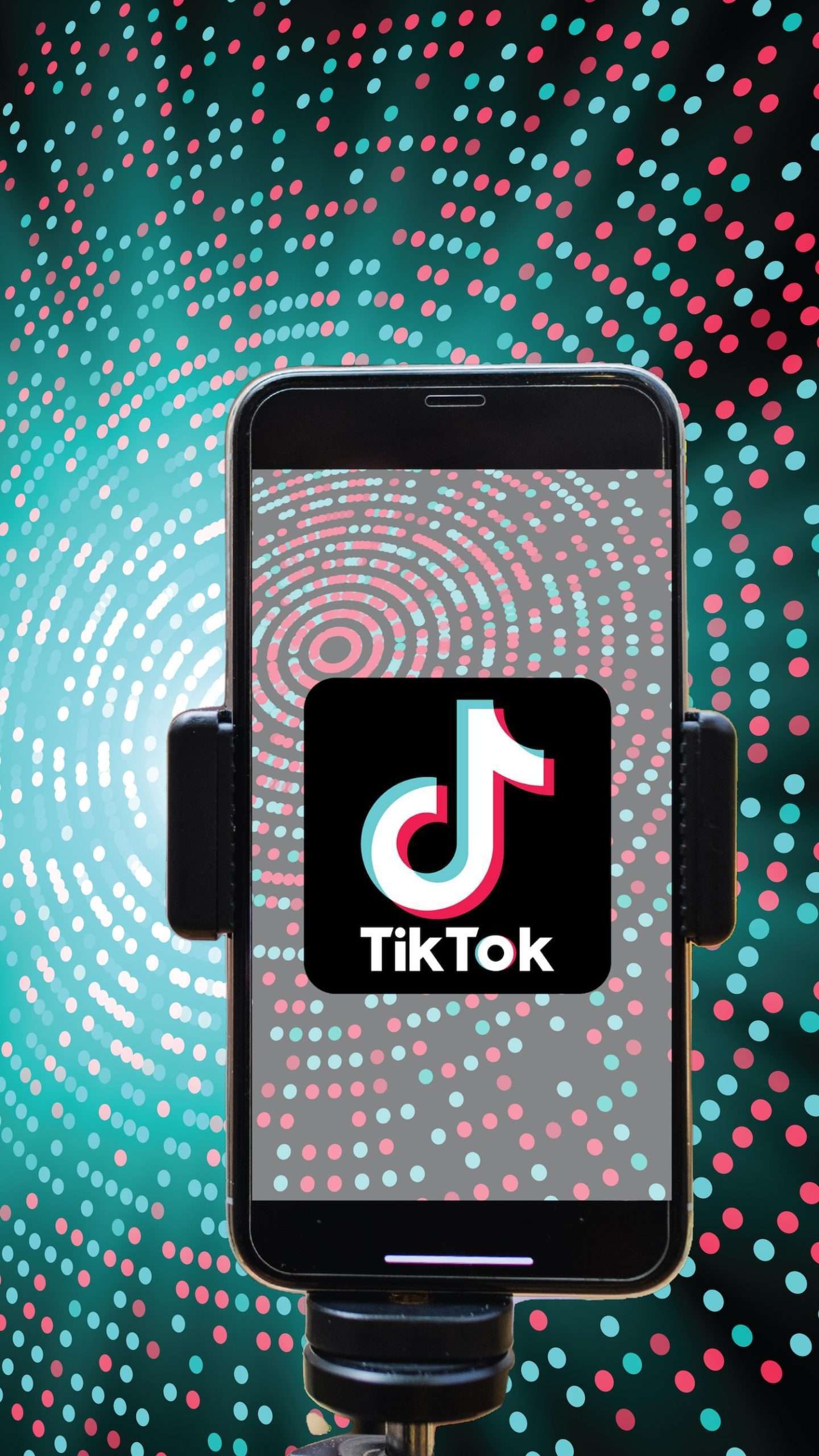 A smartphone mounted on a stand with the TikTok app logo displayed on the screen, against a backdrop with a circular dotted pattern in teal, black, and red colors.