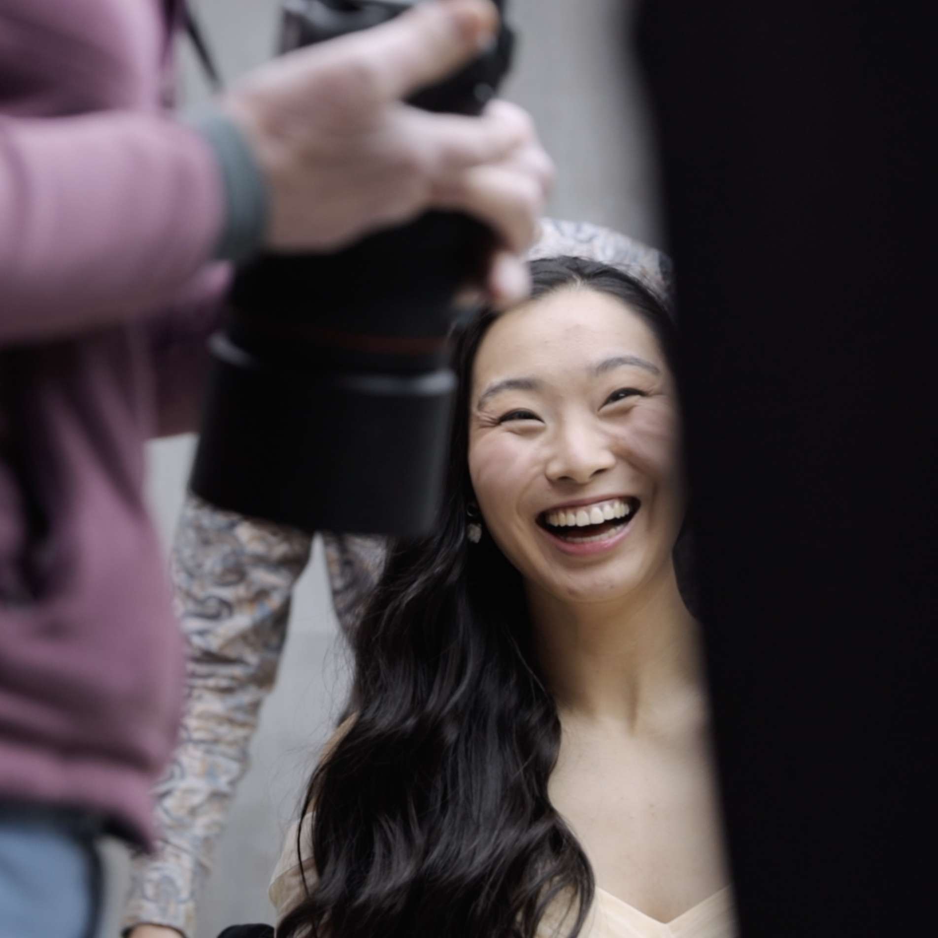 A smiling woman being photographed, seen through the edge of a camera lens with a blurry photographer in the foreground.
