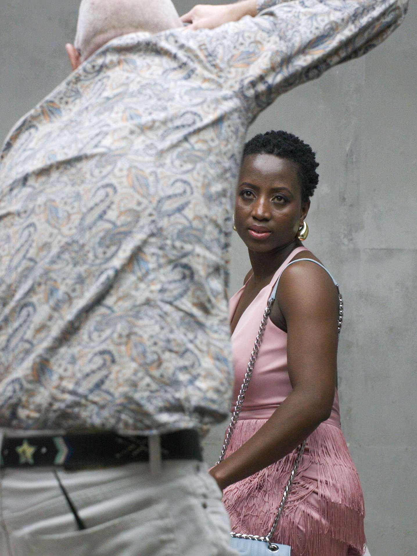 A woman in a pink sleeveless top and pleated skirt looking thoughtfully at the camera, with a partial view of a person in a patterned shirt in the foreground.
