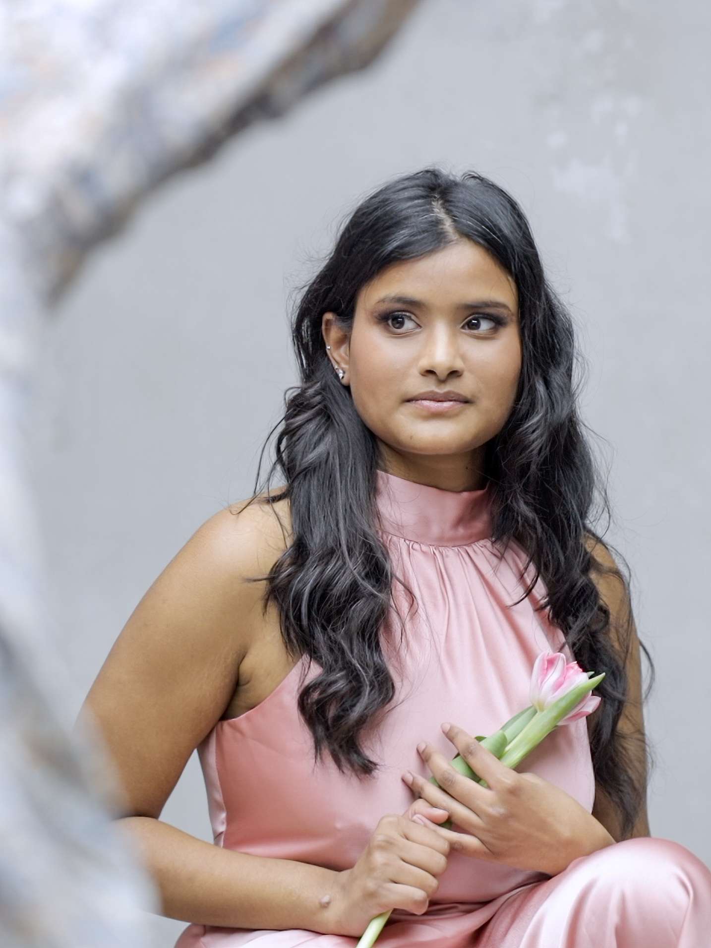 A woman in a pink dress sitting and holding a pink flower, with a thoughtful expression on her face.