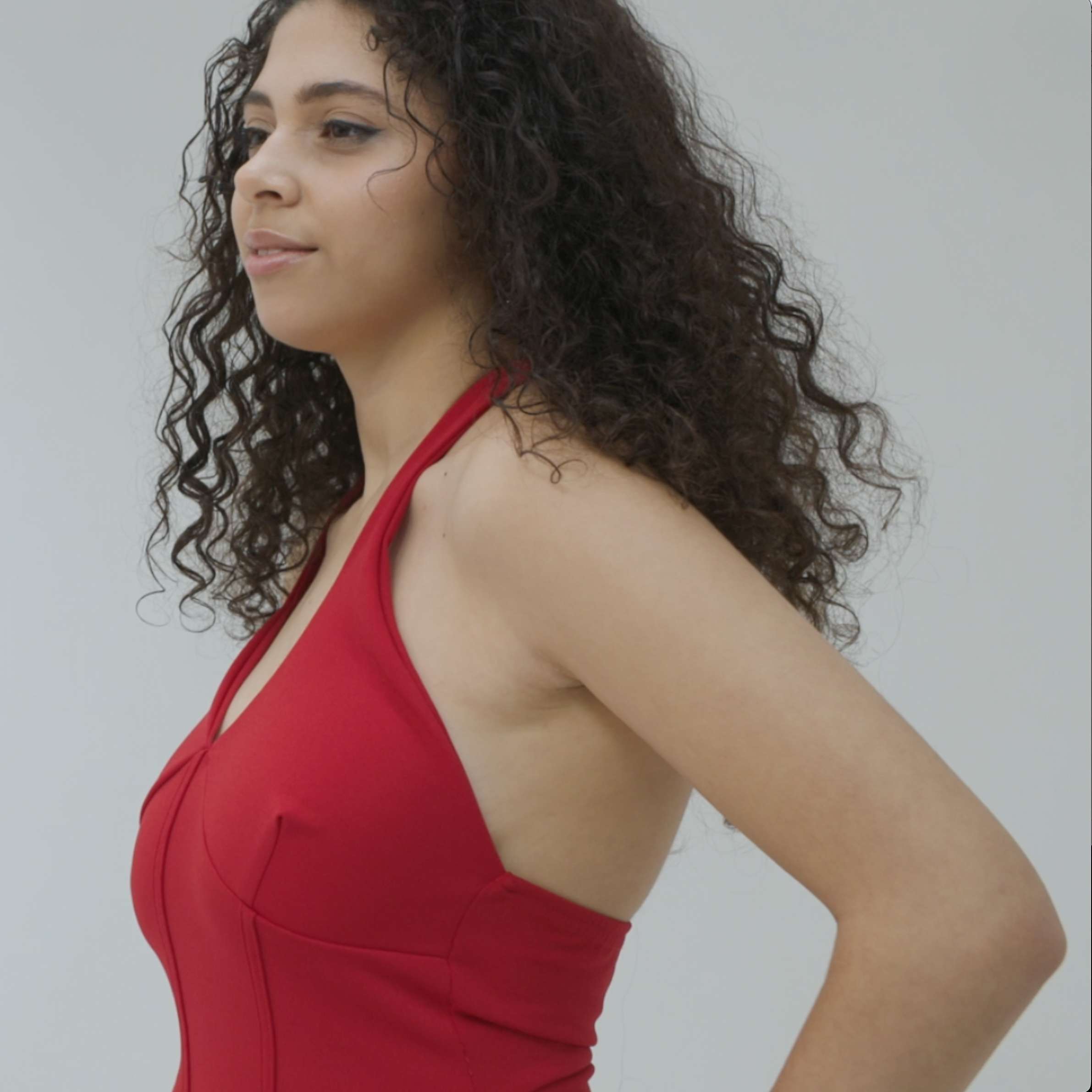 A woman with curly hair wearing a red dress, posing with a profile view against a neutral background.