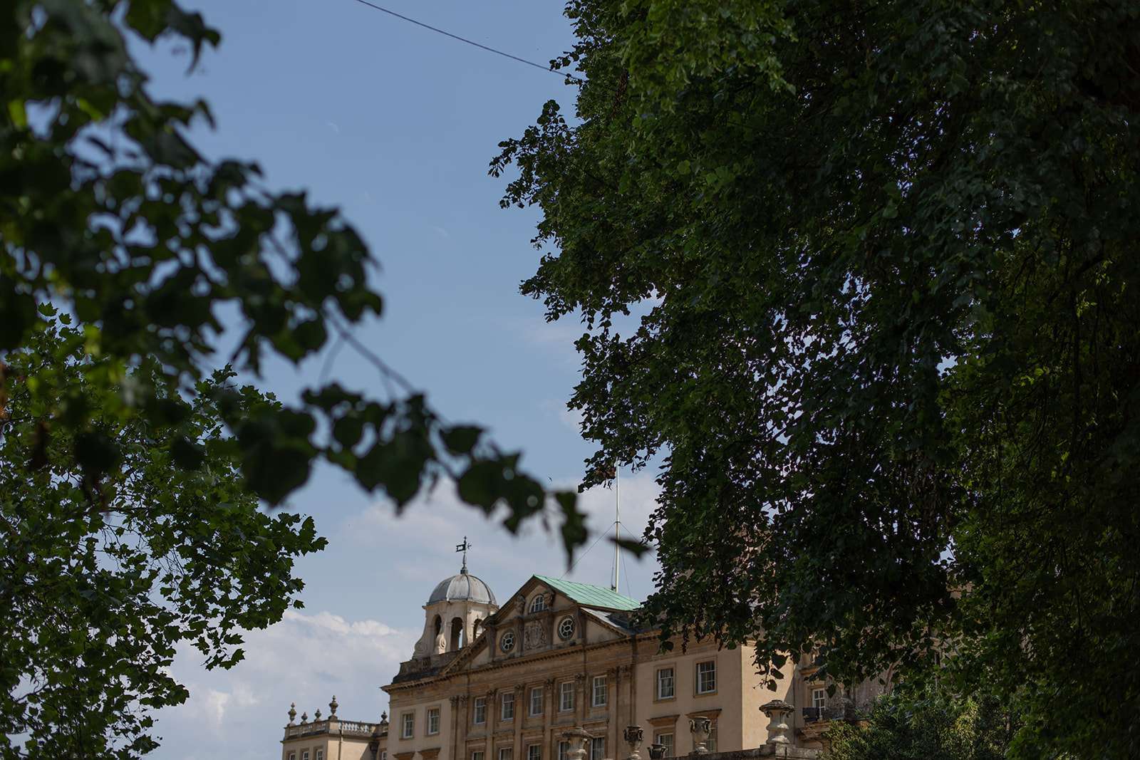 View of an old classical building with a dome, partially obscured by the leaves of green trees under a clear sky.