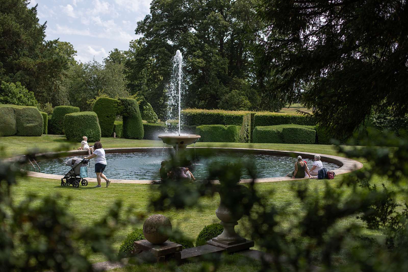 People relaxing by a water fountain in a lush garden setting with topiary and trees in the background.